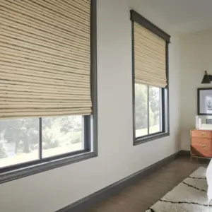 Two woven wood shades with dark wood trim in a bedroom, framing windows near a nightstand and framed art